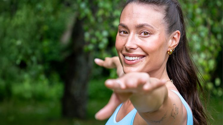 Leende kvinna i yogaposition utomhus, med grönskande bakgrund.