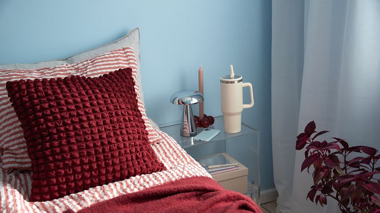 Soverom med blå vegg, stripete dynetrekk i rødt og hvitt, rød pute og pledd, nattbord i akryl med moderne kromet lampe.