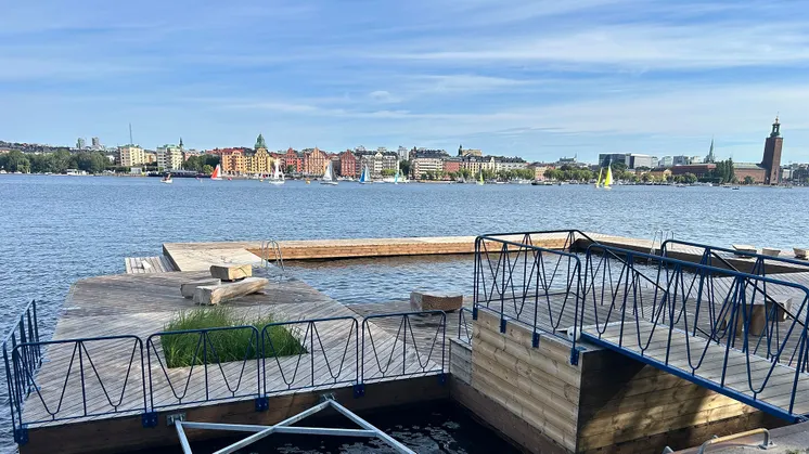 Badbryggor i vattnet vid Söder Mälarstrand i Stockholm och i bakgrunden syns båtar, stadsbilden och stadshuset i horisonten.