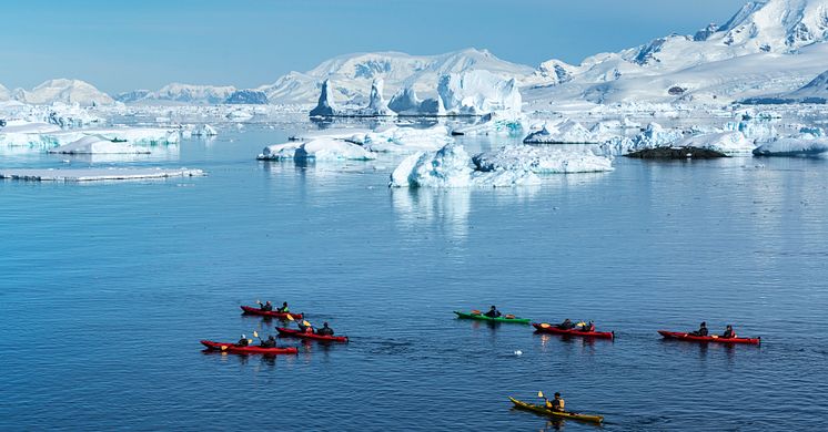 A row of kayaks cut through the icy blue waters of Antarctica with huge ice glaciers behind them, all set against a blue sky.