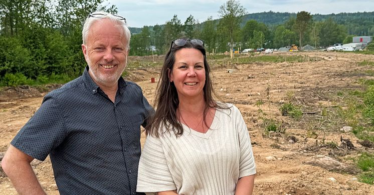 Två personer framför ett byggnadsområde där det grävs i marken. I bakgrunden syns skog och byggnader.