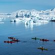 A row of kayaks cut through the icy blue waters of Antarctica with huge ice glaciers behind them, all set against a blue sky.