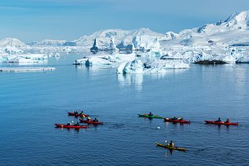 A row of kayaks cut through the icy blue waters of Antarctica with huge ice glaciers behind them, all set against a blue sky.