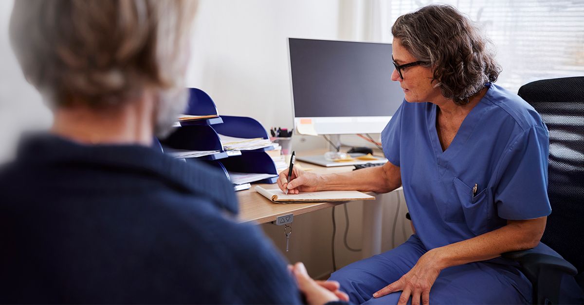 En läkare sitter vid sitt skrivbord och skriver i ett block. Mittemot sitter en patient med ryggen vänd mot kameran.