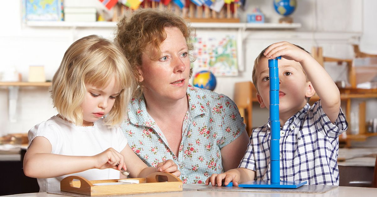 En pedagog sitter med två barn i förskolemiljö vid ett bord. Det ena barnet bygger ett torn av blåa runda klossar och det andra barnet pysslar på en träbricka.