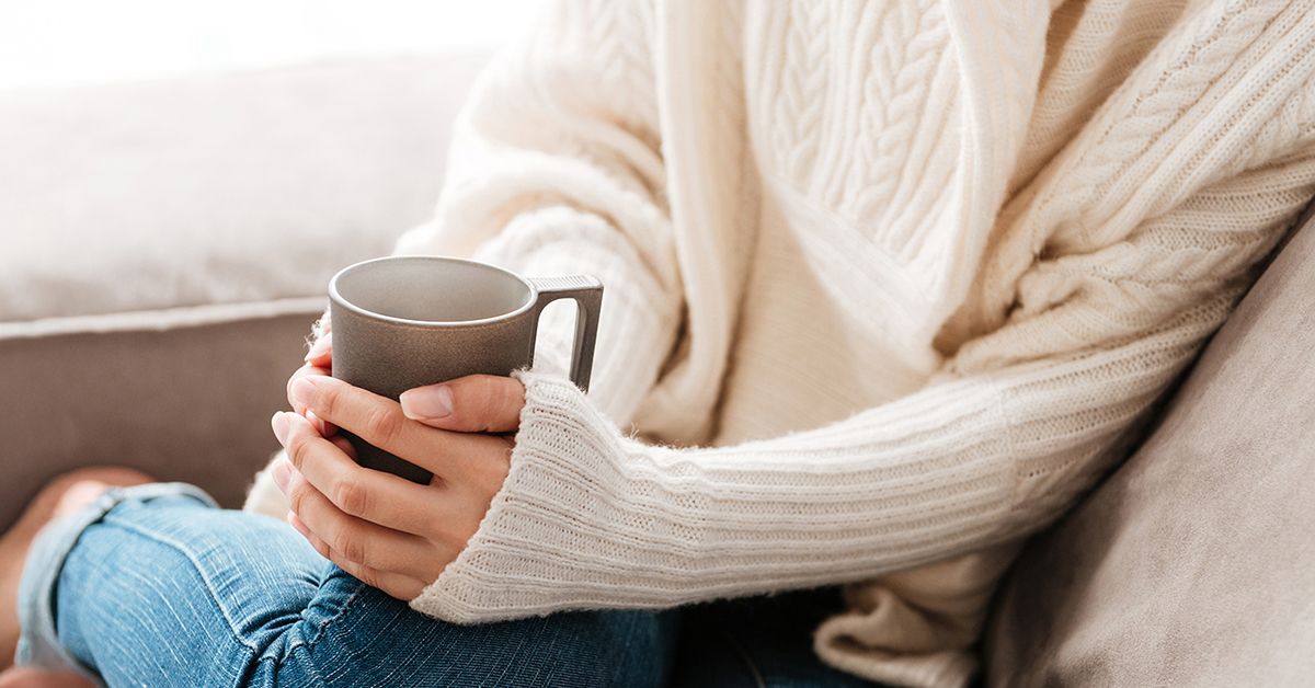 En person i en vit stickad tröja och blå jeans sitter uppkrupen i en beige soffa och håller händerna kupade runt en kaffekopp.
