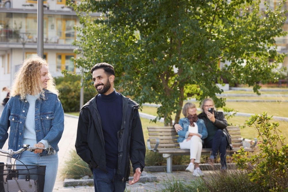 Syntolkning: Ett par på promenad, äldre par på en parkbänk i bakgrunden