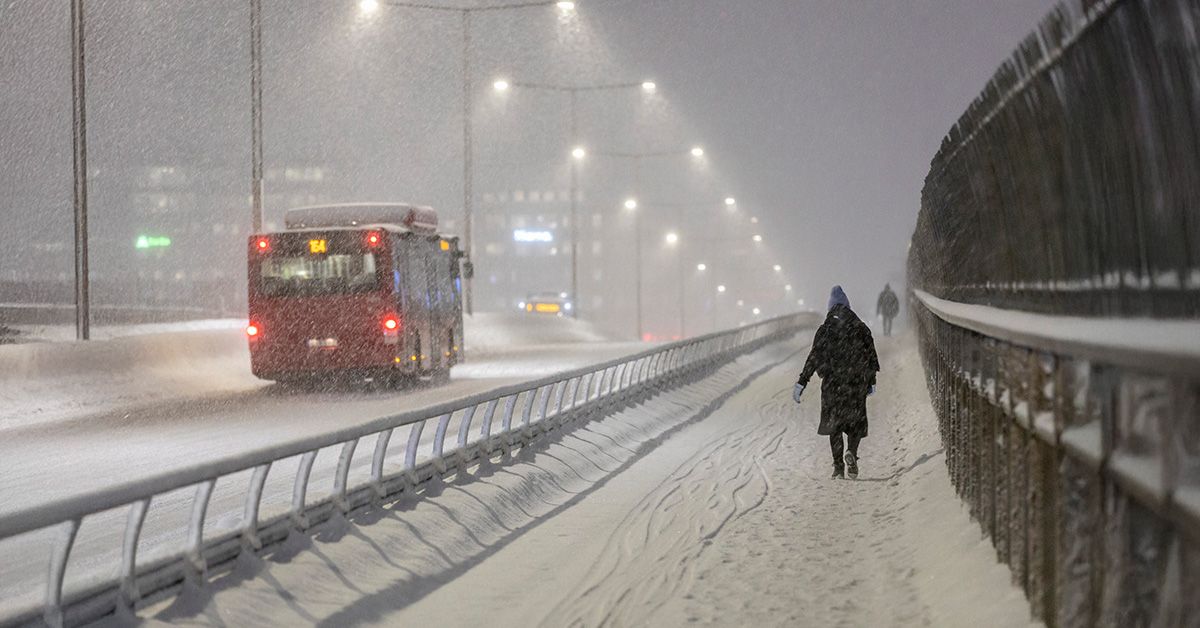 Snöig väg där en röd buss passerar en kvinna som går på trottoaren.
