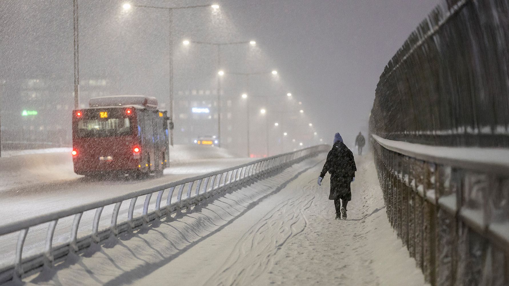 Snöig väg där en röd buss passerar en kvinna som går på trottoaren.