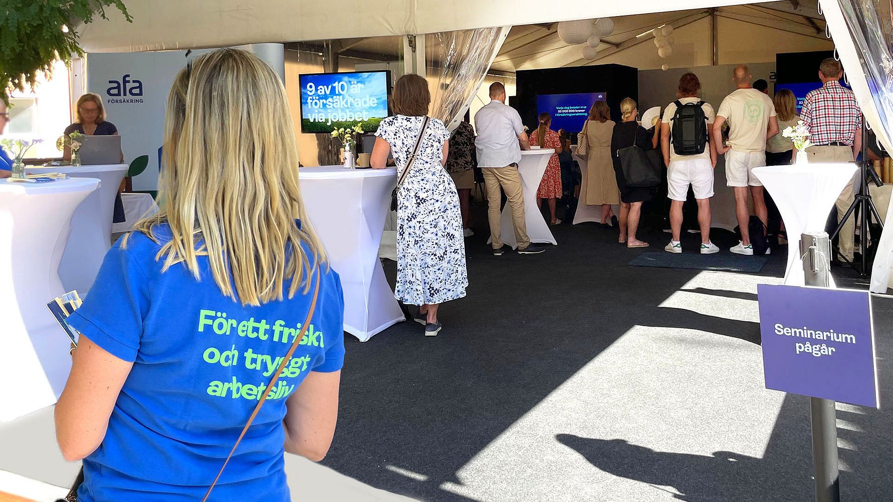 Seminarium pågår på Afa Försäkrings arena i Almedalen. I förgrunden syns en person med t-shirt med texten För ett friskt och tryggt arbetsliv och i bakgrunden pågår ett seminarium inne i ett stort tält med många i publiken.