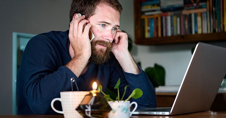 En man med skägg sitter vid ett bord i hemmiljö framför en dator och pratar i telefon. På bordet står ett ljus och en tekopp.