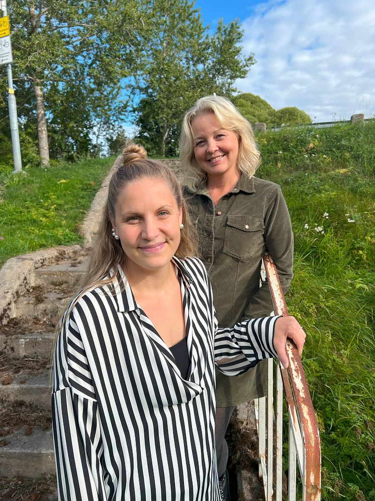 Matilda och Camilla står i nedre delen av en utomhus trapp. I bakgrunden syns del av ett broräcke.
