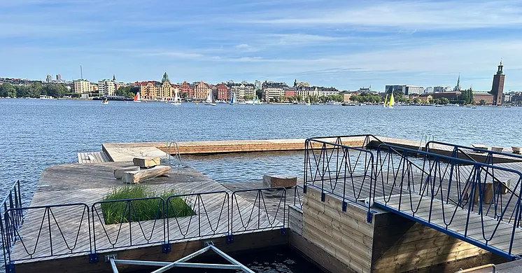 Badbryggor i vattnet vid Söder Mälarstrand i Stockholm och i bakgrunden syns båtar, stadsbilden och stadshuset i horisonten.