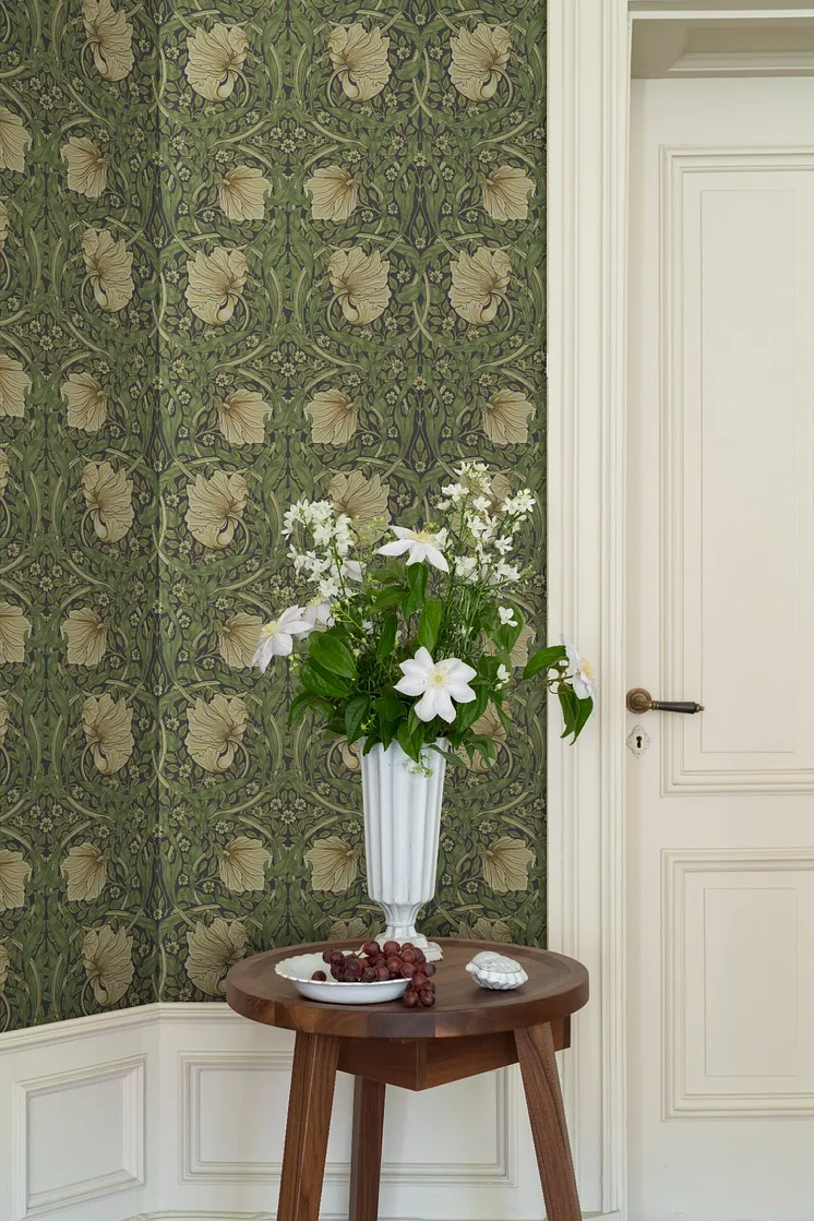 Detaljrik blommönstrad tapet i gröna och beige toner, vit spegeldörr och träbord med vas fylld av vita blommor.
