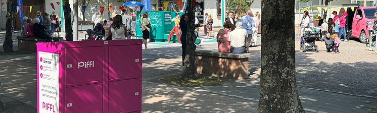  En rosa Piffl-aktivitetsbox står vid en park där människor samlas. Träd kastar skuggor över boxen, och i bakgrunden syns bän