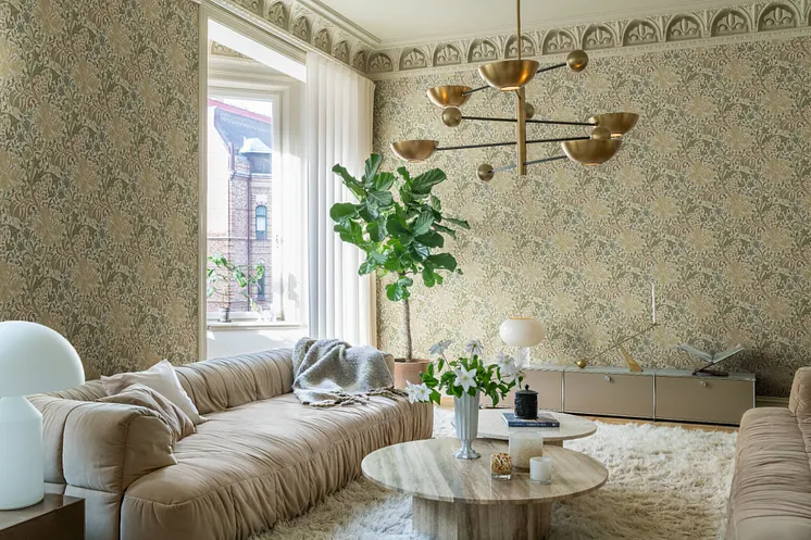 Ljust vardagsrum med detaljrik blommönstrad tapet i beige och dämpade gröna toner, mjuka soffor och mässingsfärgad taklampa.