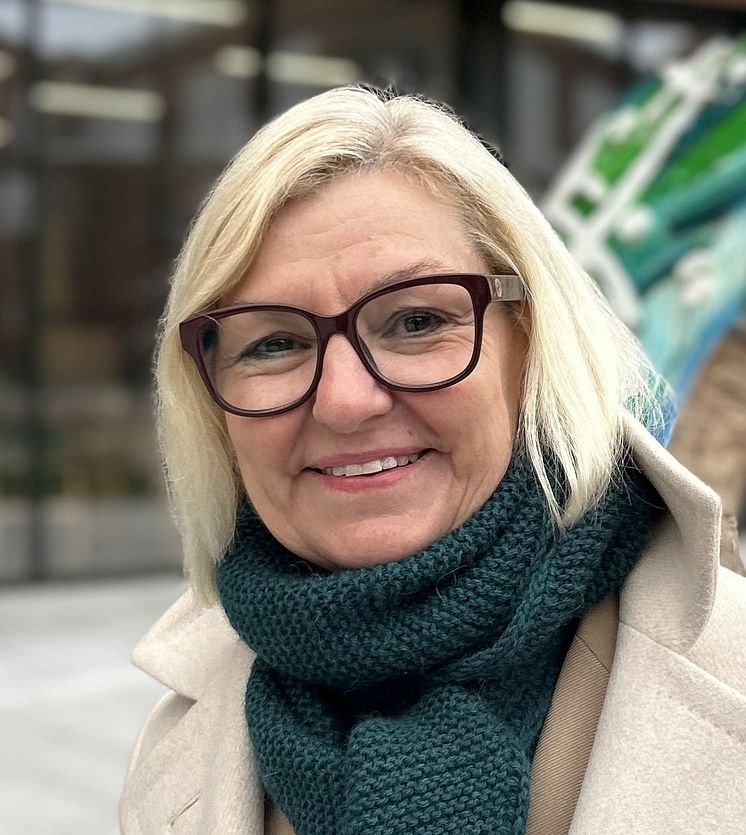 Bilde av Eli Grimsby, en blond kvinne med briller, skjerf og beige kåpe, i urbant miljø.
