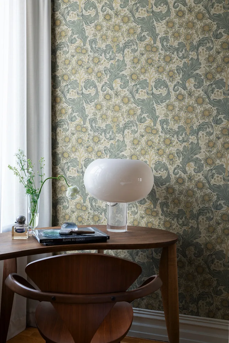 Ljus blommönstrad tapet med gula blommor och slingrande bladverk i blågröna toner, träbord och vit bordslampa.