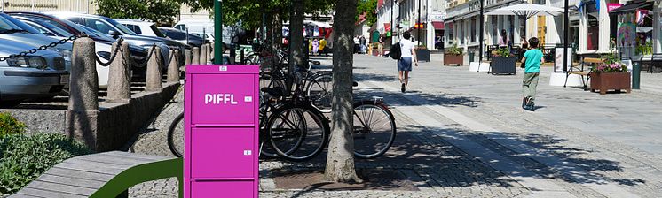 En rosa Piffl-box placerad längs en gågata i centrala Alingsås. I bakgrunden syns människor som promenerar.