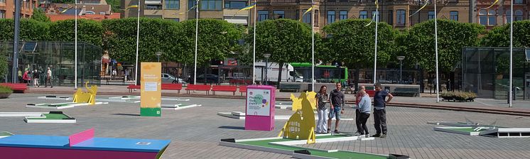 Pifflbox på Sundstorget i Helsingborg med människor som spelar minigolf runt omkring, solig dag och grönskande träd i bakgrun