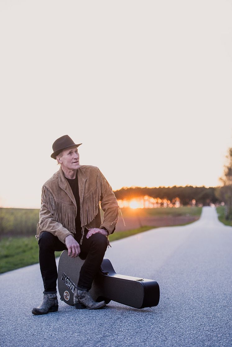 Lasse Sigfridsson sitter på ett hårt gitarrfodral i mitten av en landsväg. Han är iklädd hatt, jacka och boots. Foto.