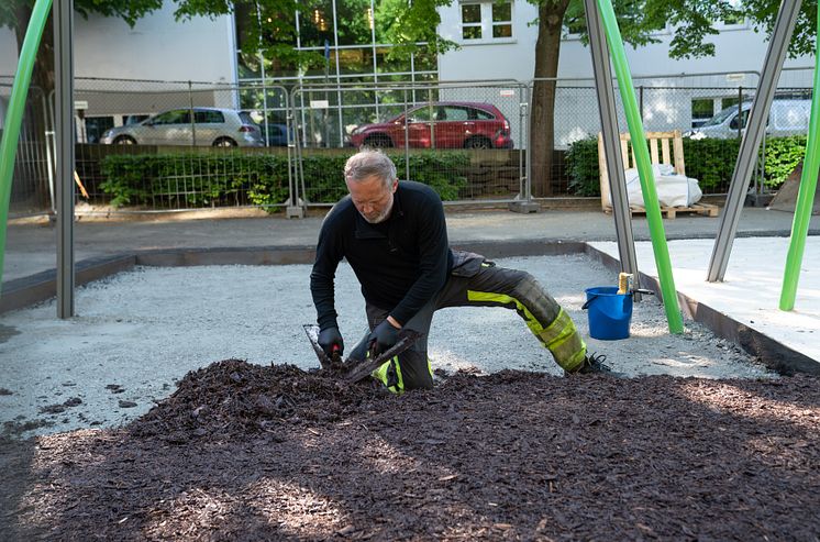 Person sprer mørk bark med verktøy på lekeplass, iført arbeidsklær med refleks.