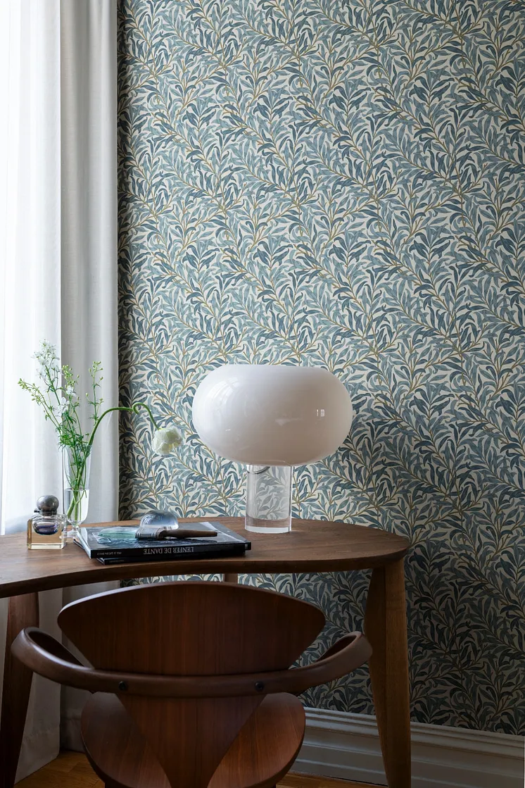 Blå bladmönstrad tapet med slingrande grenar, träbord, designlampa och dekorativa detaljer.
