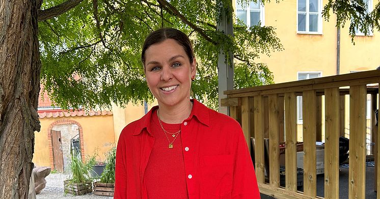 Lina Ejlertsson står under ett lövträd och ser in i kameran. Hon har på sig en röd tröja, röd skjorta och vita byxor.
