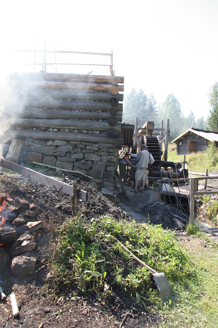 Besökare framför utslagsbröstet. Rostning av malm pågår i rostbåset. I bakgrunden till höger en av åtta färskningssmedjor. Från järnförsöket på Lapphyttan 2008. Foto: Eva Hjärthner-Holldar.