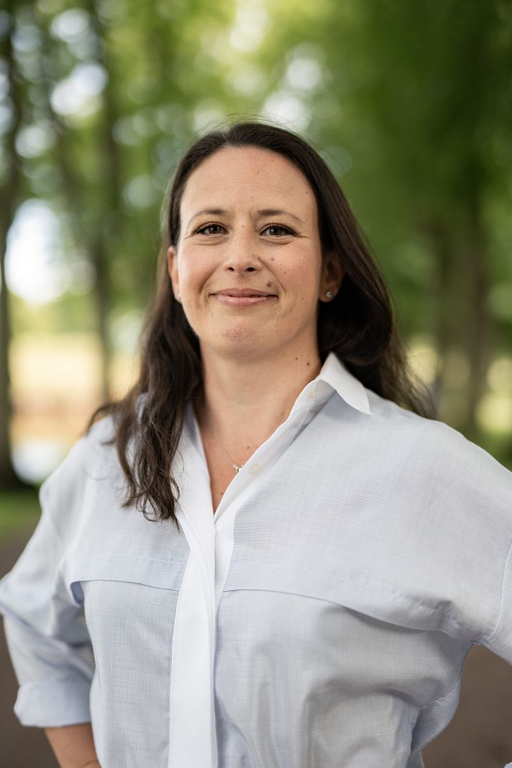 Leende kvinna i vit blus som står i en grön allé.