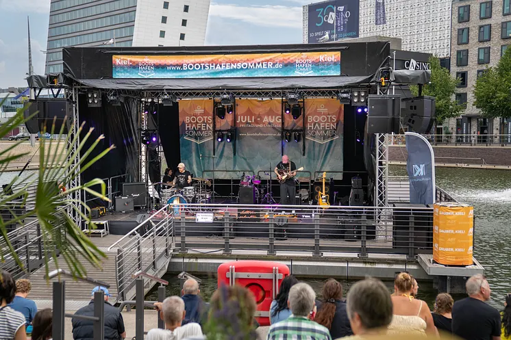 Eine Band steht auf der Bühne des Bootshafensommers. Menschen schauen und hören zu.