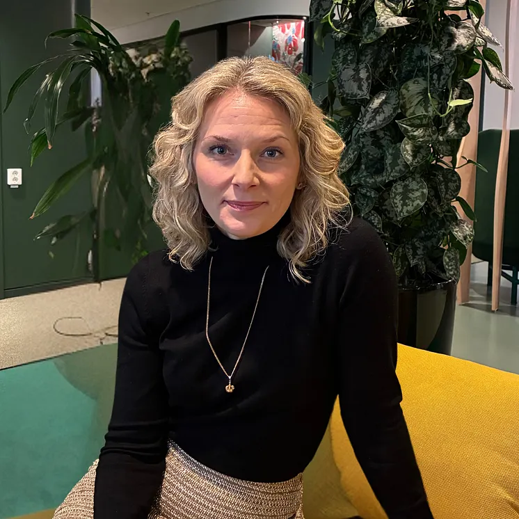 Anna Williamsson har ljust, utsläppt hår, svart tröja och ett halsband hon sitter i en gul soffa framför en stor grön växt. 