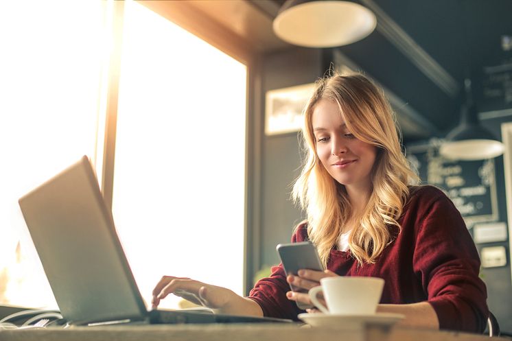 Ung kvinne på café som jobber på både mobil og laptop