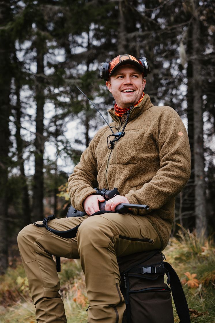  Leende jägare i fleecejacka från Pinewood sitter på jaktväska i skogen med gevär i knät.