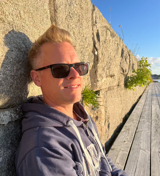 Herman Saming sitter mot en stenmur en solig dag. Han har en hoodie och solglasögon på sig. Foto. 
