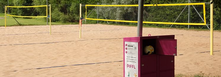 Pifflbox vid beachvolleyplan med nät och boll, omgiven av grönska i ett somrigt landskap i Linköping.  En volleyboll syns.