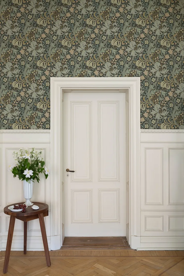 Klassisk interiör med detaljrik blommönstrad tapet i dämpade gröna och blå toner, vit bröstpanel och spegeldörr.