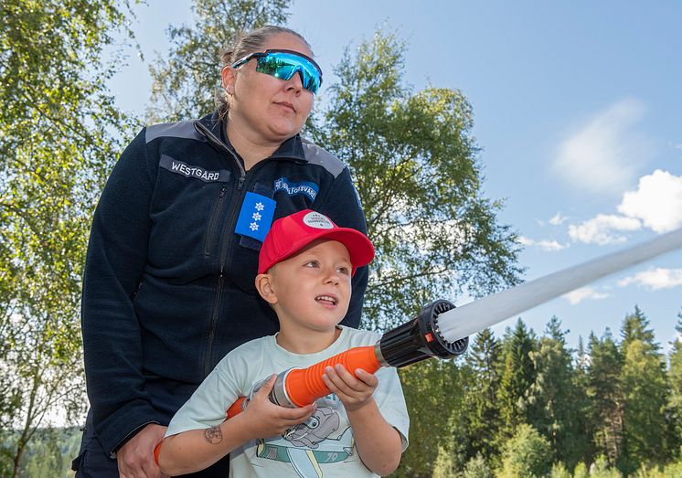 En kvinnelig offiser fra Sivilforsvaret hjelper en liten gutt å spyle med brannslange.