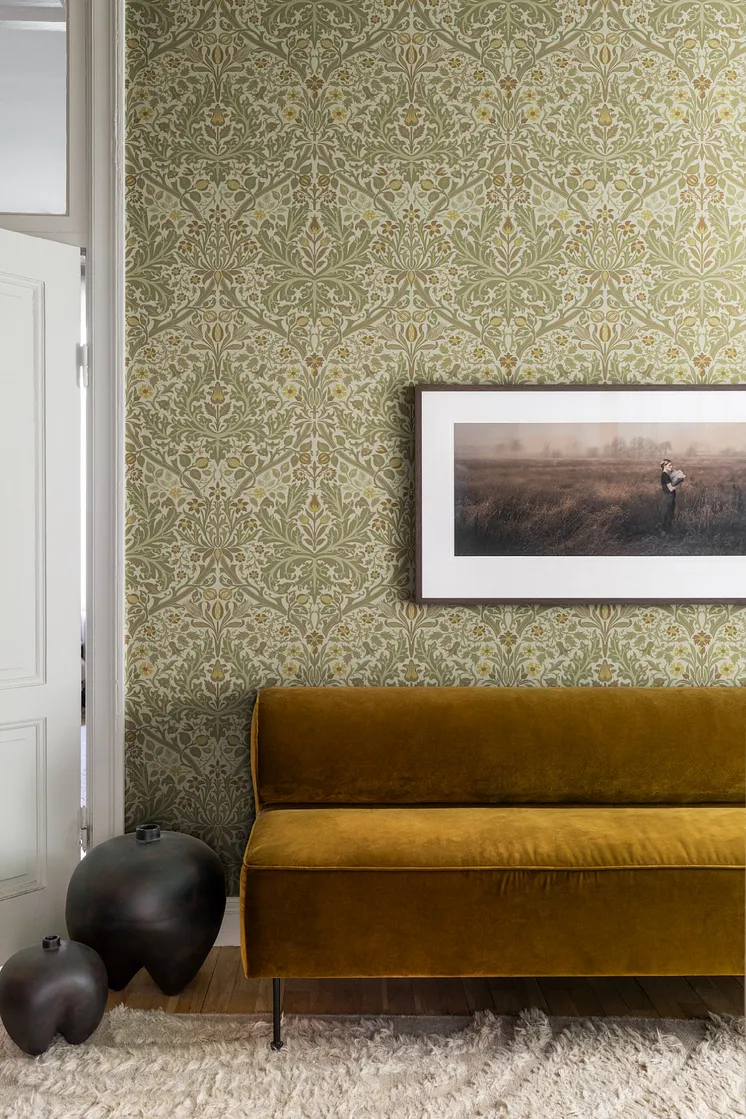 Interiör med ljus botanisk tapet i nougat, khaki och beige, senapsgul sammetssoffa och inramad tavla.