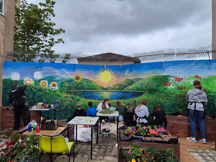 Green hills and sunset mural painted on a brick wall with people working on the mural and garden in front.