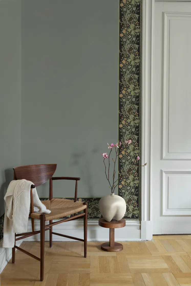 Gråblå vägg med dekorativ blommönstrad tapetbård runt dörr och golvlist, trästol och vas med blommor.