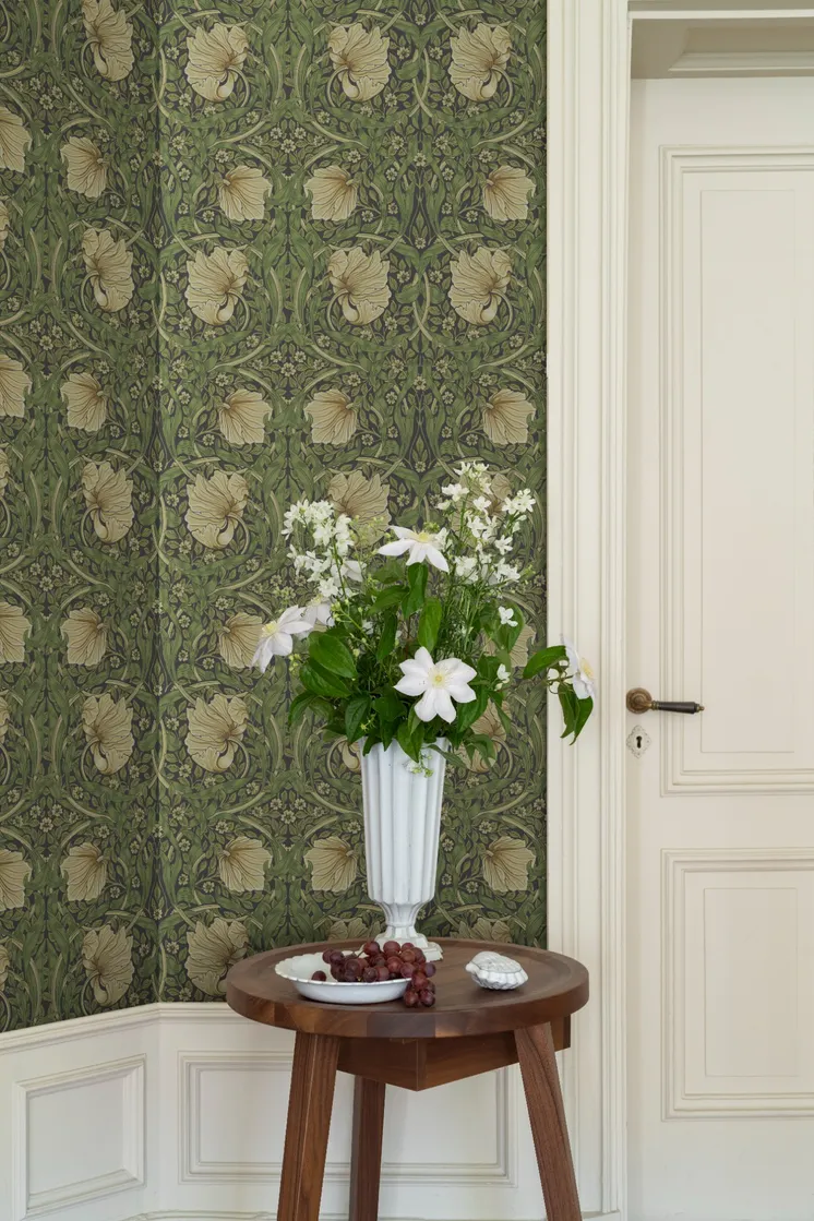 Detaljrik blommönstrad tapet i gröna och beige toner, vit spegeldörr och träbord med vas fylld av vita blommor.