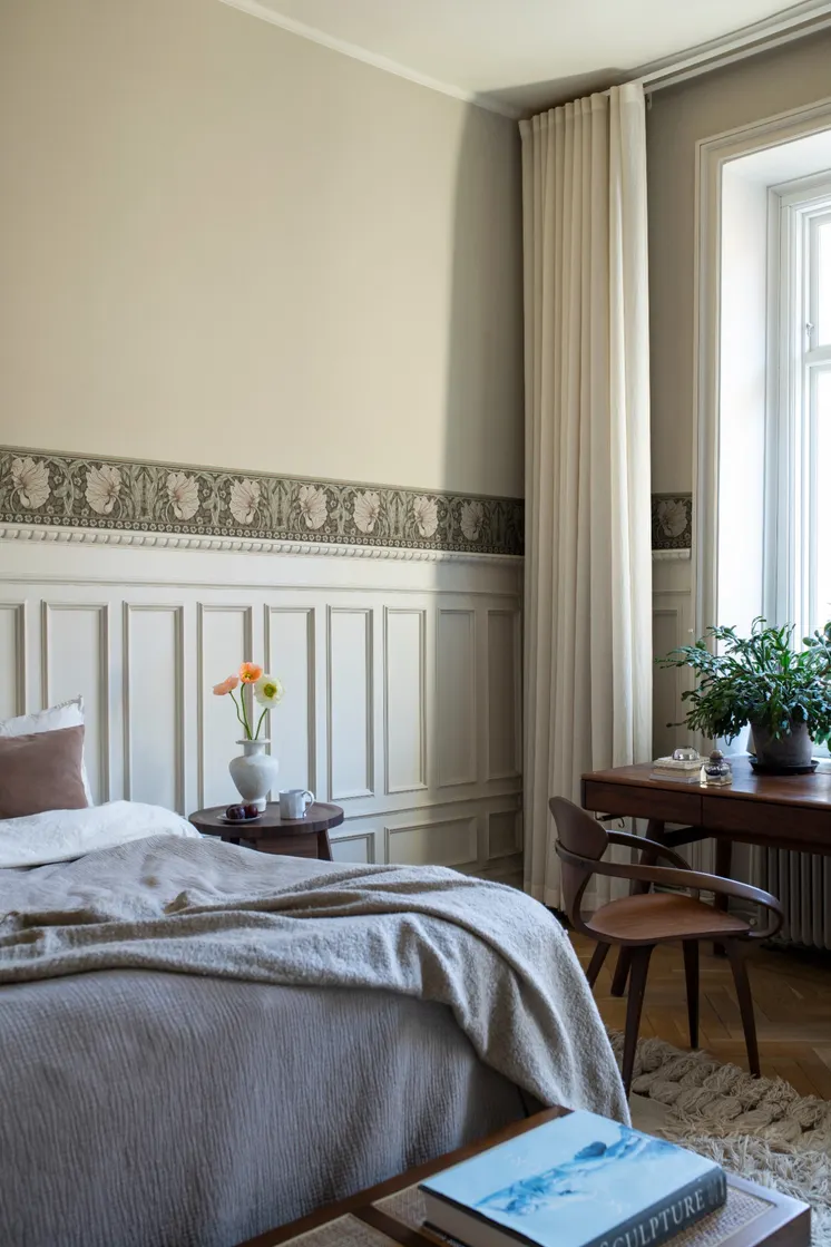 Ljust sovrum med dekorativ blommönstrad tapetbård, beige väggar, beige bröstpanel och trädetaljer.