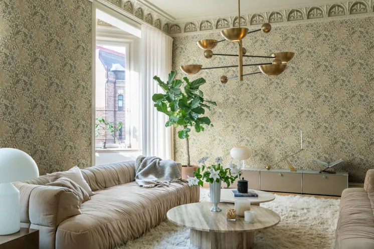 Ljust vardagsrum med detaljrik blommönstrad tapet i beige och dämpade gröna toner, mjuka soffor och mässingsfärgad taklampa.