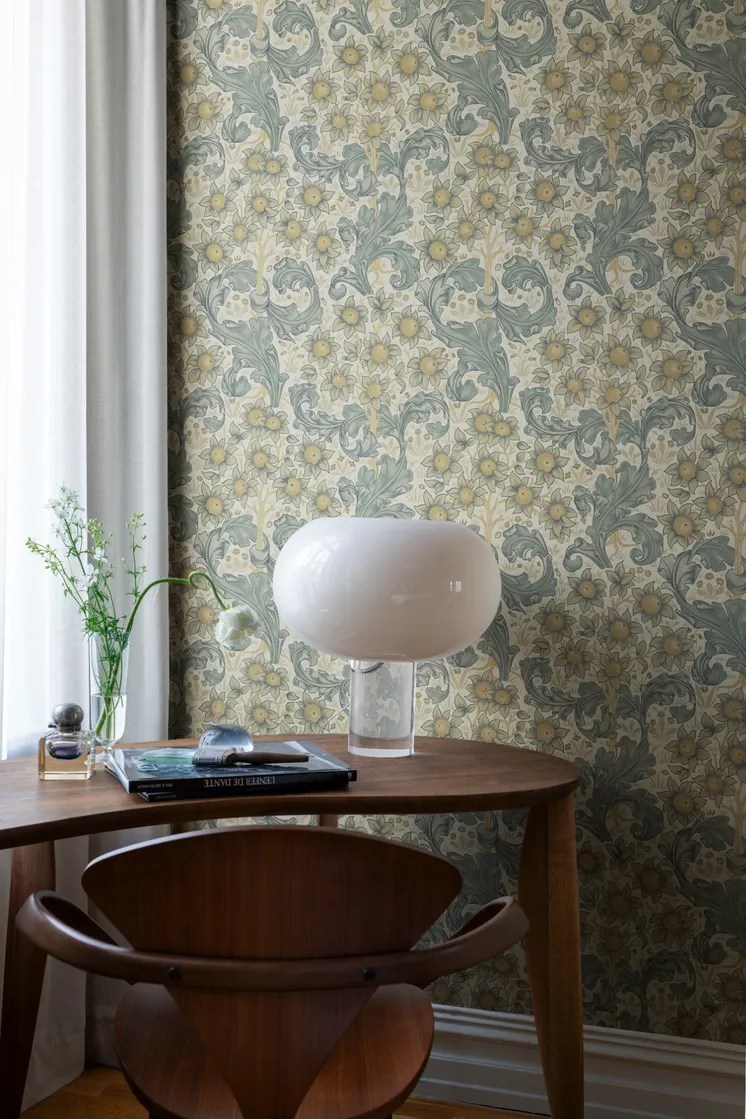 Ljus blommönstrad tapet med gula blommor och slingrande bladverk i blågröna toner, träbord och vit bordslampa.