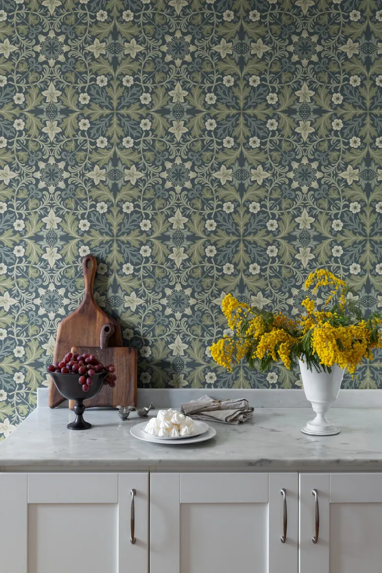 Kök med blågrön blommönstrad tapet, marmorbänk, träskärbrädor och vas med gula blommor.