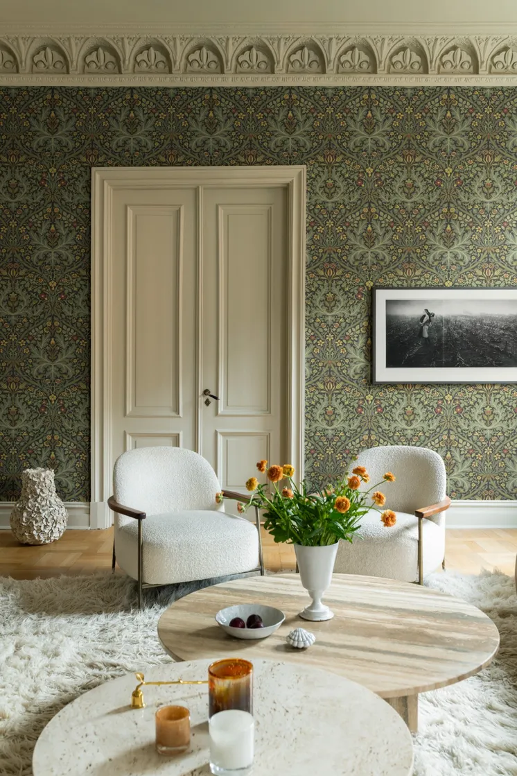 Vardagsrum med detaljrik botanisk tapet i dämpade gröna och varma toner, ljusa fåtöljer, marmorbord och orange blommor.