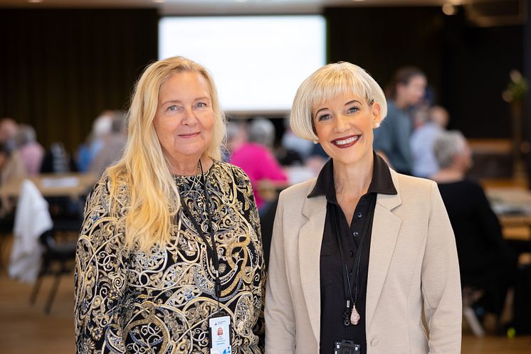 Två leende kvinnor poserar i ett stort mötesrum, i bakgrunden ses många stående och sittande deltagare på evenemanget.