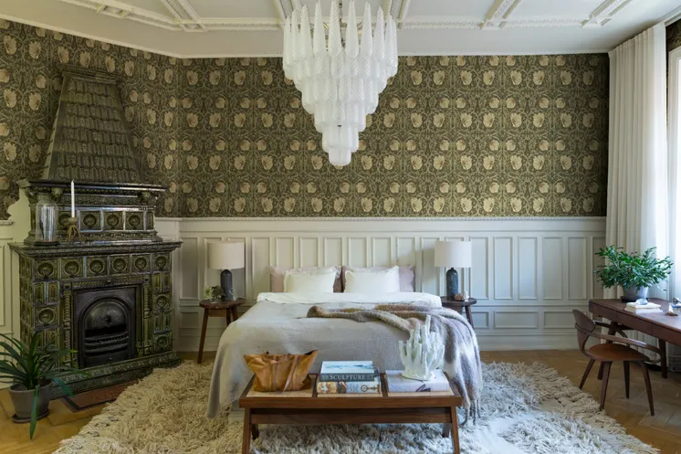 Rymligt sovrum med detaljrik blommönstrad tapet i dämpade gröna och beige toner, vit bröstpanel och kakelugn