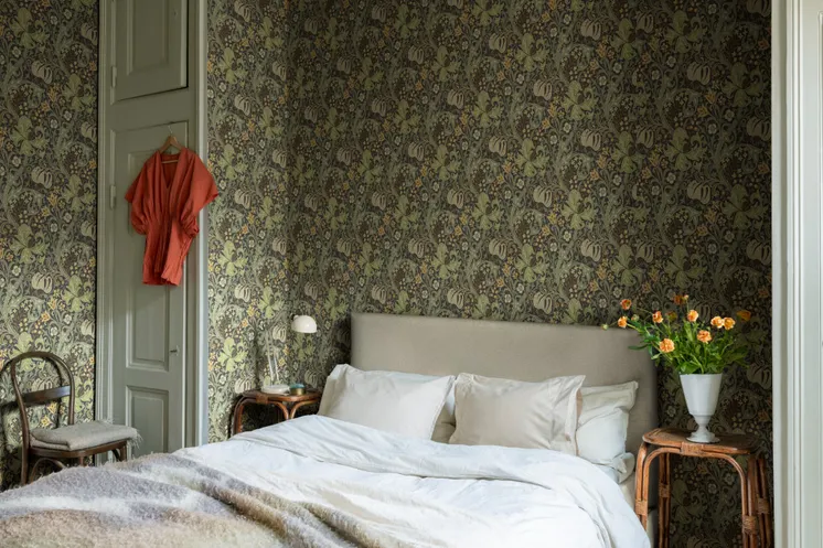 Sovrum med mörk, detaljrikt blommönstrad tapet i gröna toner, ljus säng och rottingmöbler.